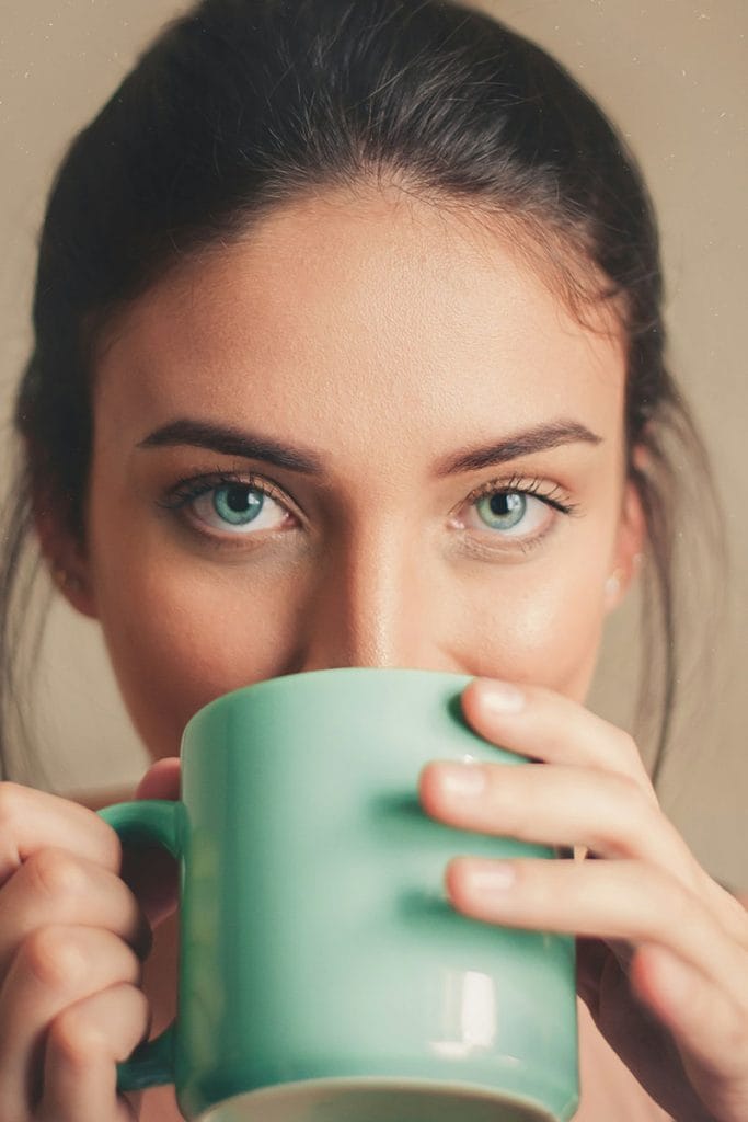 Beautiful woman with striking blue-green eyes, sipping coffee from a blue-green mug, showcasing the rejuvenated look achievable with blepharoplasty by Dr. Randall in Morristown, NJ.